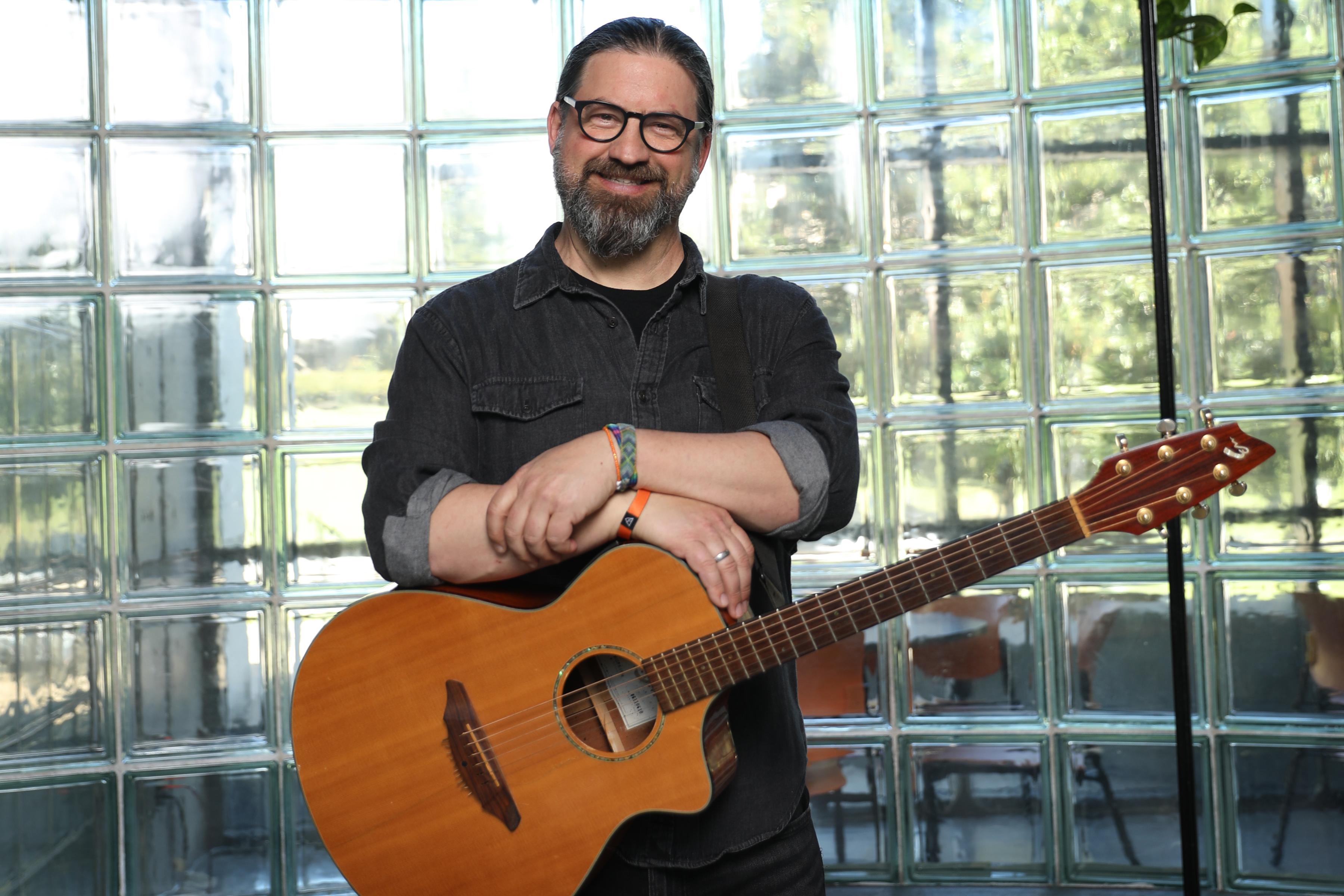 Jason LeVasseur - Smiling with guitar in front of glass wall.